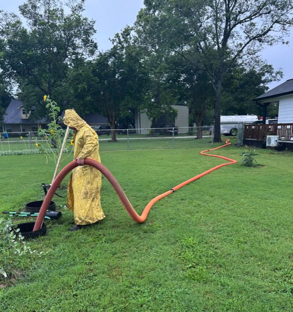 A OnePump worker in a rain slicker holding the suction hose over an open septic tank, hose running across the lawn to the truck